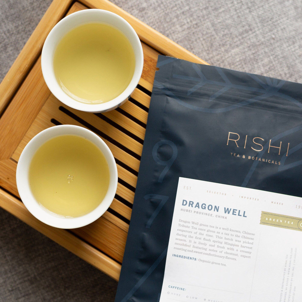 A cup of Dragon Well tea and a bag of Dragon Well tea on a wooden tray. (Brand: Rishi Tea & Botanicals)