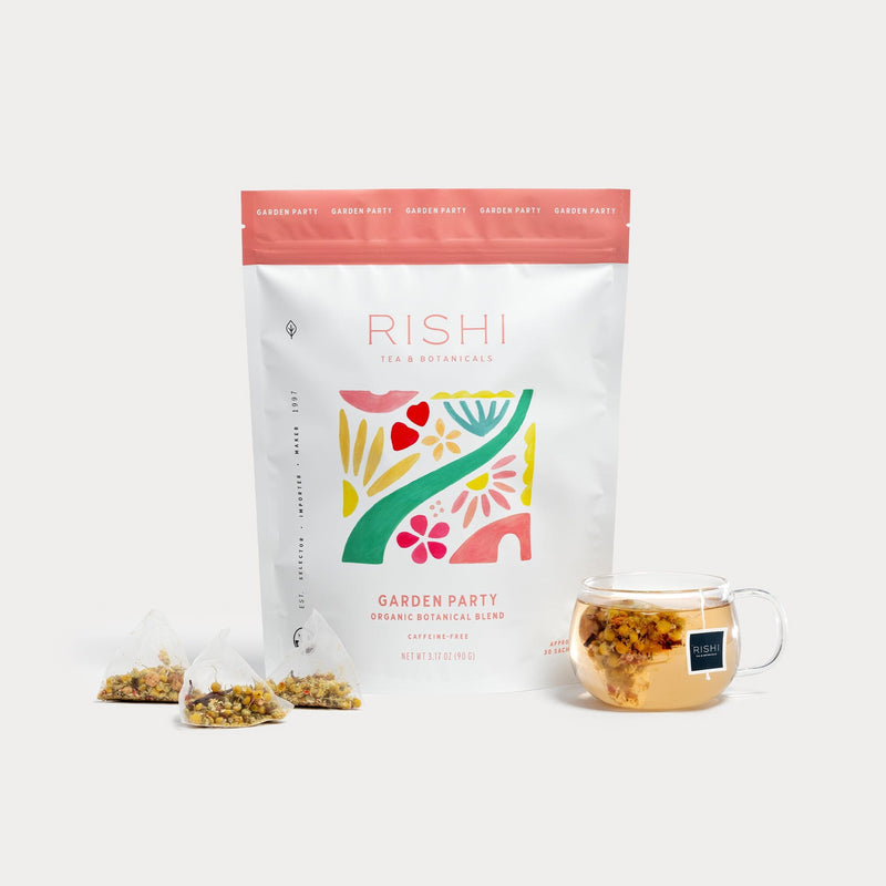 A Rishi Tea & Botanicals Garden Party organic botanical tea blend, with chamomile and honeybush, three pyramid sachets, and a glass cup of brewed tea with botanicals are displayed on a white background.