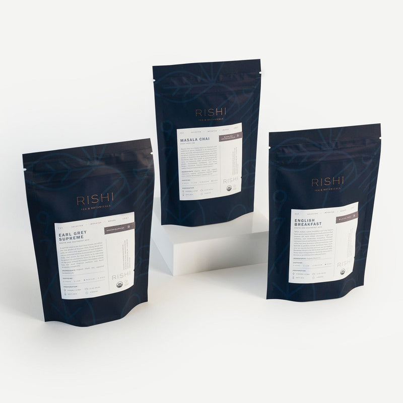 Three stand-up pouches of Rishi Tea & Botanicals—energizing blends including Earl Grey Supreme, Masala Chai, and English Breakfast—are displayed upright, with one pouch elevated on a white block.