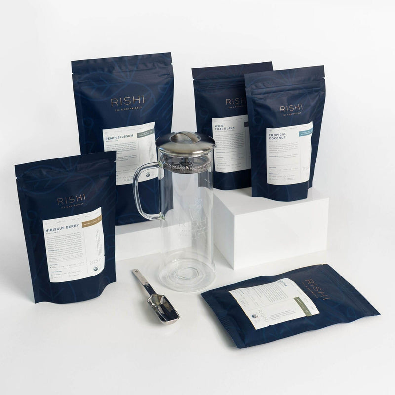 Five navy blue packets of Rishi Tea & Botanicals, a clear glass pitcher with metal lid, a metal tea scoop, and an iced tea bundle are displayed on a white surface.