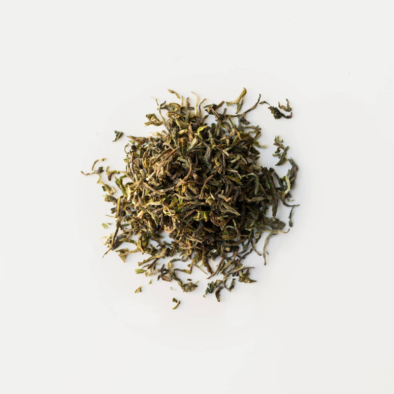 A small pile of Darjeeling First Flush Chamong EX2 by Rishi Tea & Botanicals on a white background. The dried loose-leaf tea showcases green and brown hues, with subtle hints of stone fruits interwoven among the fragrant blossoms.