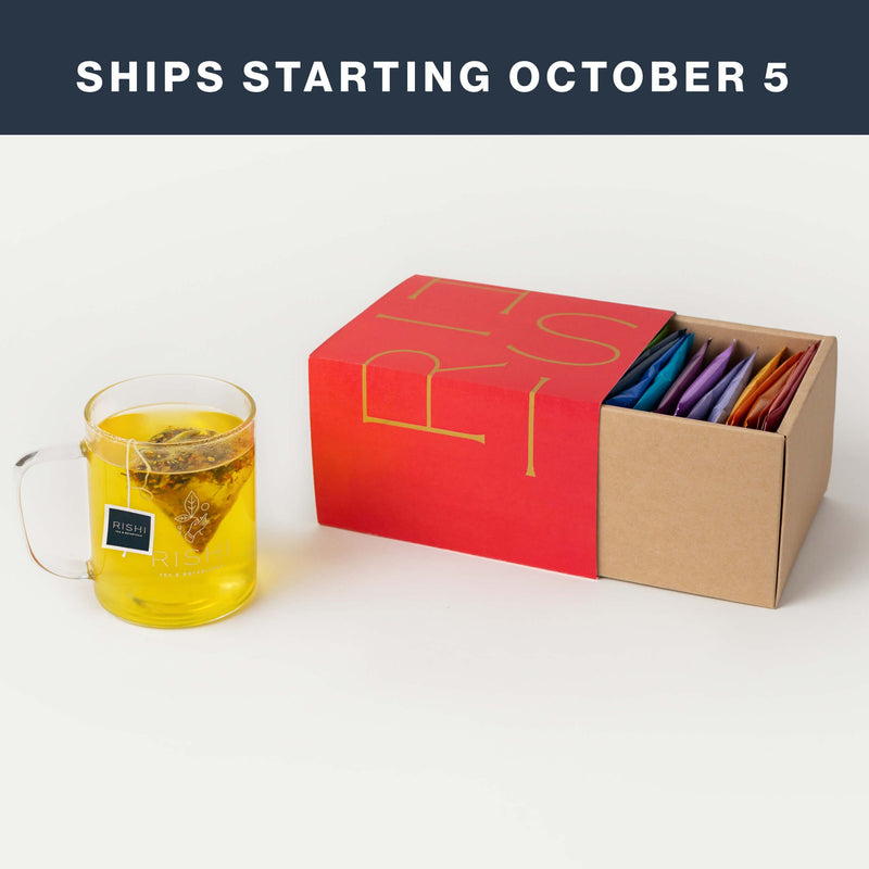 A glass mug of tea sits next to an open red and brown box containing assorted tea sachets from a curated teas collection. Text above reads, “SHIPS STARTING OCTOBER 5.”.