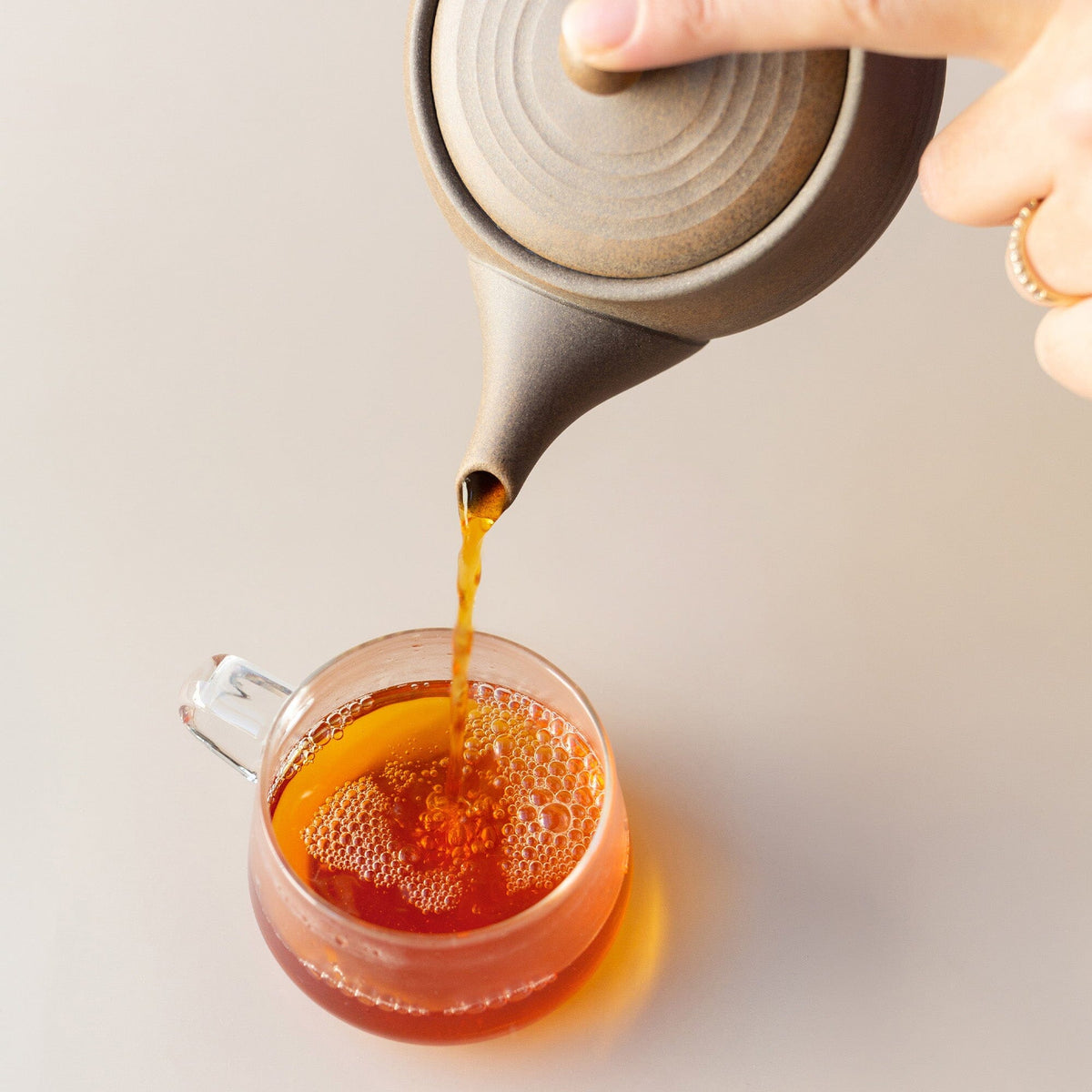 A hand pours tea from a Chato Co., Ltd. Tokoname kyusu made of shudei clay into a clear glass cup, showcasing the traditional ceramesh filter and the rich amber hue of the tea.