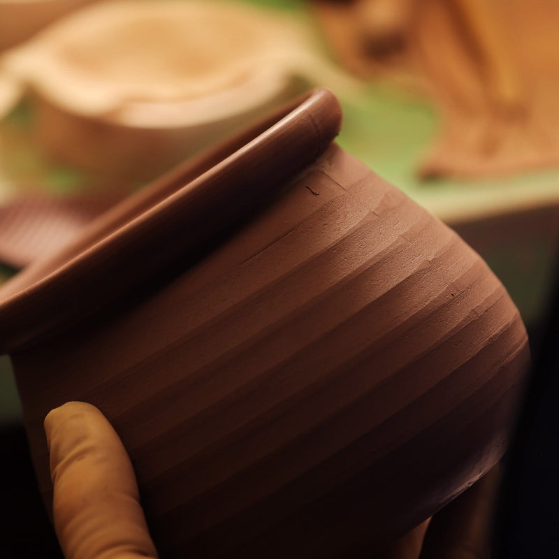 A gloved hand holds an unfinished, ribbed shudei clay pot with a smooth rim by Chato Co., Ltd. in a pottery workshop setting.