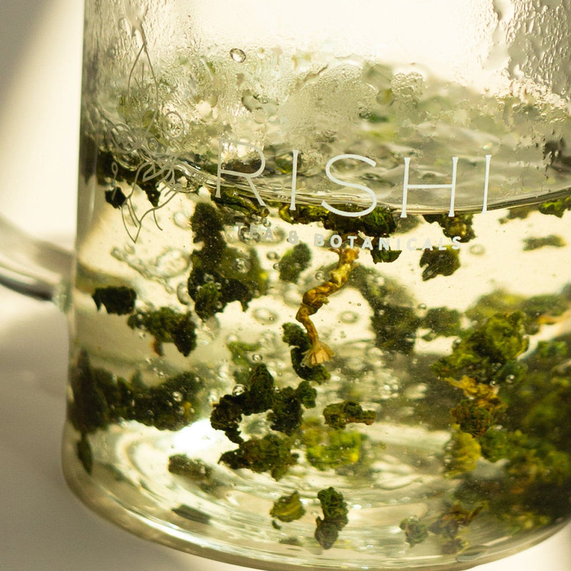 Loose leaf tea steeping in hot water inside a clear glass mug labeled 