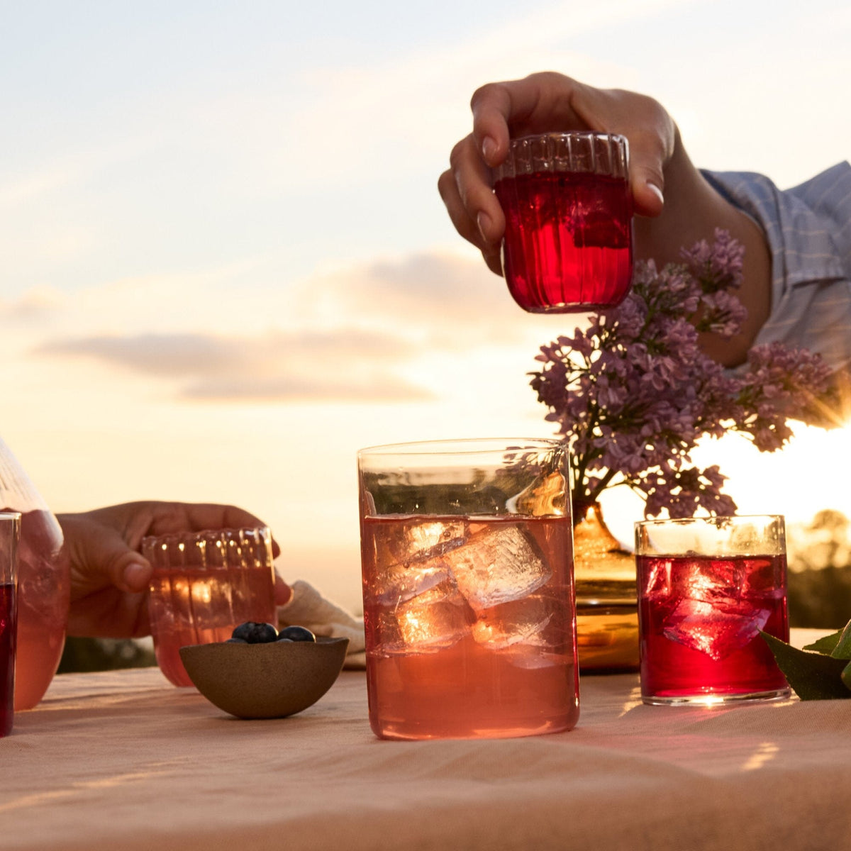 Two people hold glasses of pink and red Rishi Tea & Botanicals drinks with ice, infused with fresh herbs, placed on a table alongside a bowl of blueberries and flowers outdoors at sunset.