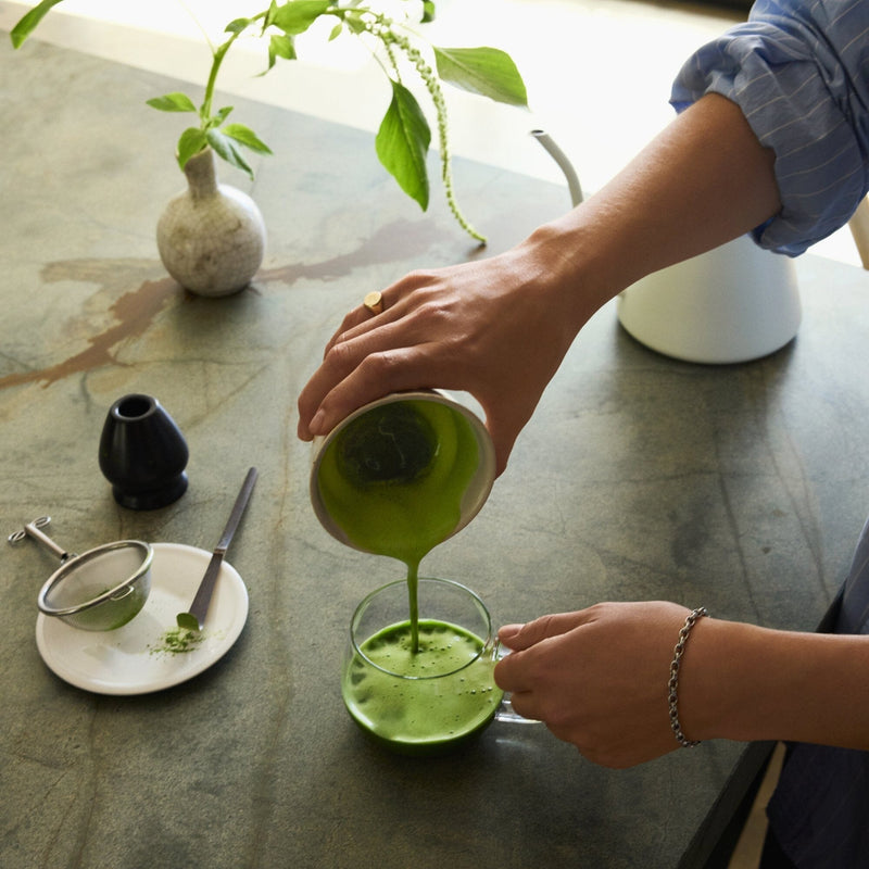 A person pours vibrant green Rishi Tea & Botanicals organic matcha tea from a small pitcher into a glass cup on a kitchen counter, with a strainer and a small plate nearby.