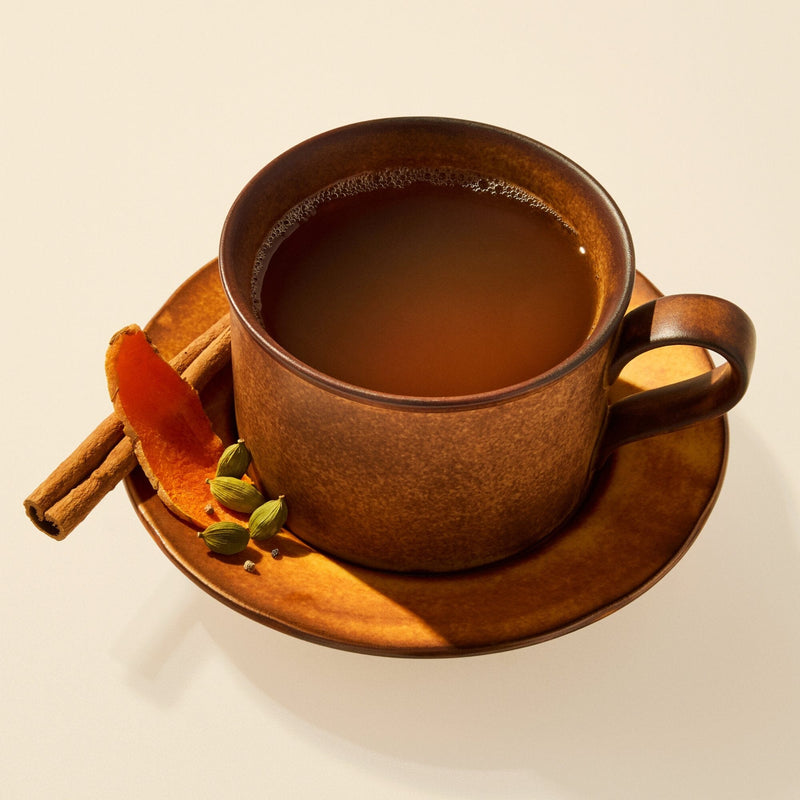 A brown ceramic cup and saucer filled with Rishi Tea & Botanicals caffeine-free chai, garnished with a cinnamon stick, green cardamom pods, and an orange peel on the side.