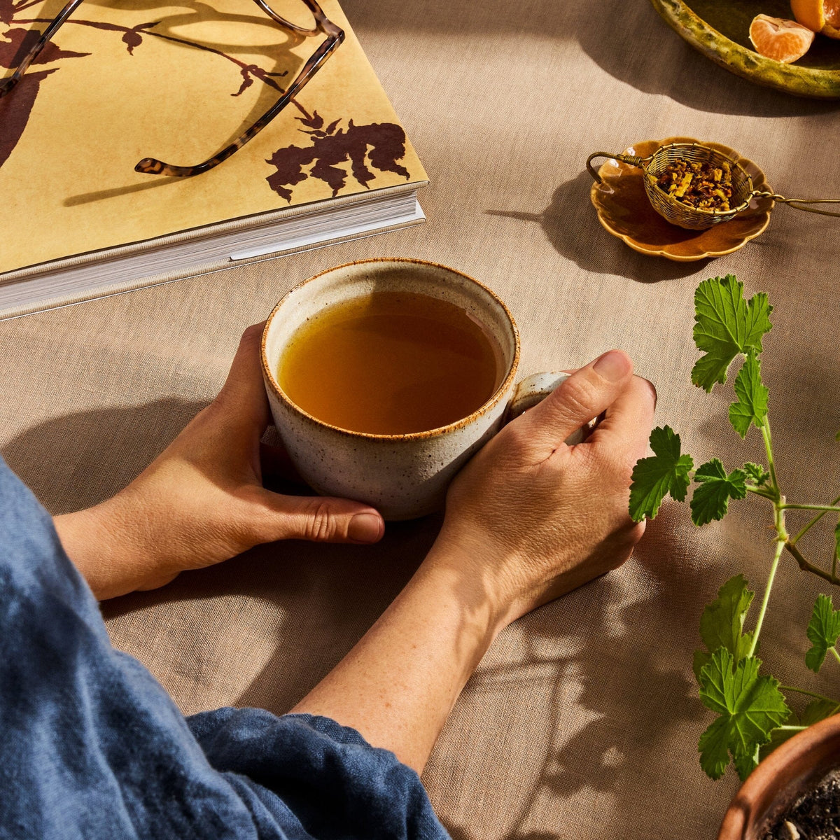 A person holds a cup of tea at a beige table alongside a Rishi Tea & Botanicals notebook, glasses, a small dish of turmeric-infused loose tea, and some greenery.
